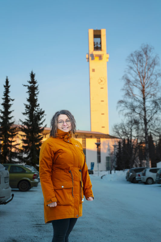 Talvisessa kuvassa on hymyilevä Heli Lavila päällään oranssi toppatakki, taustalla lunta,  Lakeuden Risti -kirkko ja kirkkotorni. 