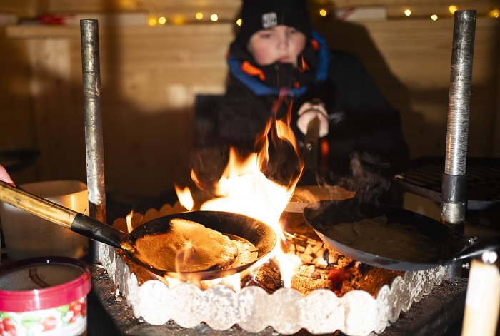 Avotulella useampi valurautainen lettupannu. Tummiin pukeutunut hahmo näkyy taustalla.