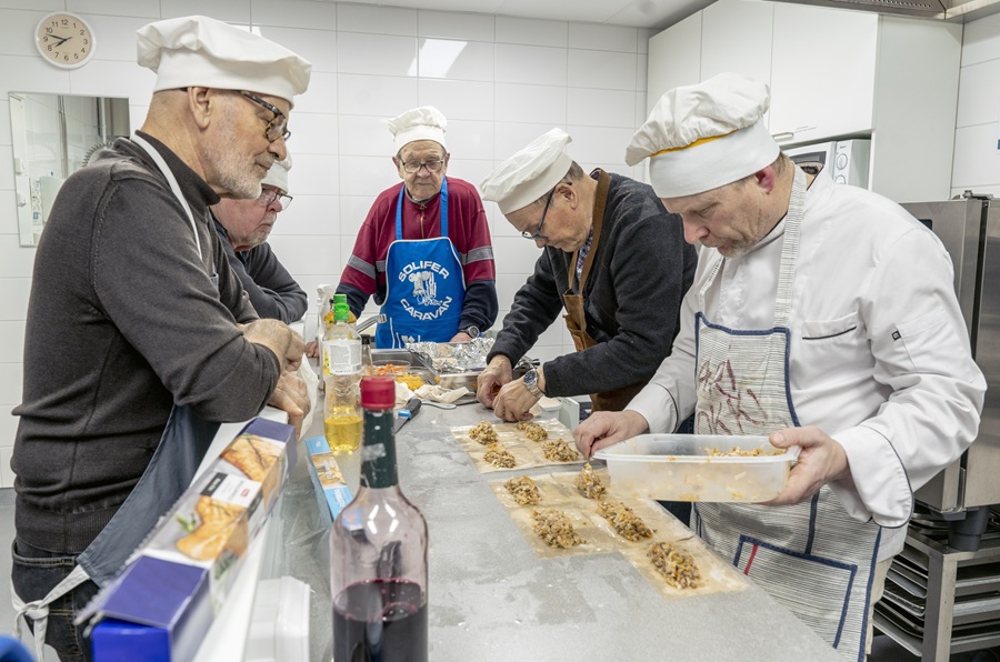 Viisi kokkihattuista iäkkäämpää miestä keittiössä tekemässä ruokaa.